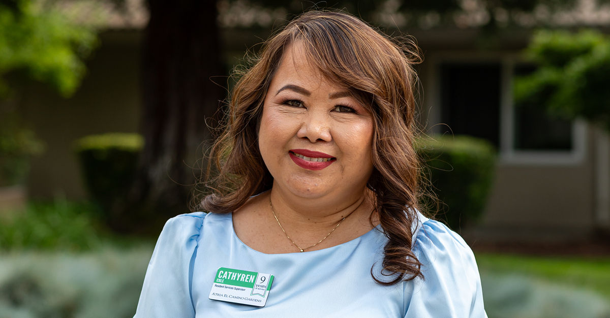 Smiling woman with long hair and a name tag, wearing a light blue blouse, standing outdoors with greenery in the background.