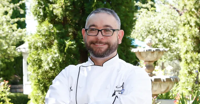 A smiling man with glasses and a beard wearing a chef's coat stands outdoors, with greenery and a fountain in the background.