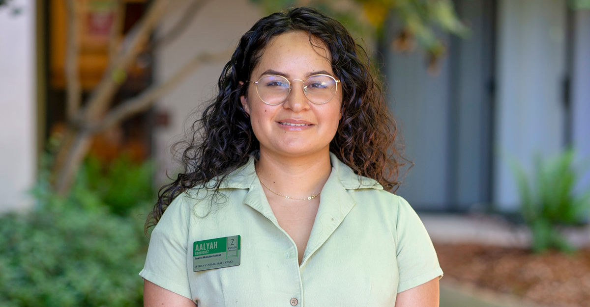 A person with long curly hair and glasses, wearing a light green uniform with a name tag, stands outside with greenery in the background.