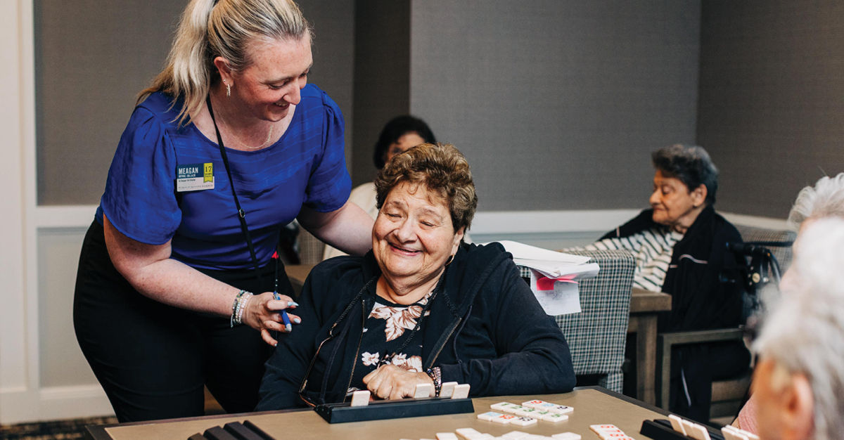 A caregiver smiles and supports an older woman seated at a table, surrounded by dominoes, in a communal living space.
