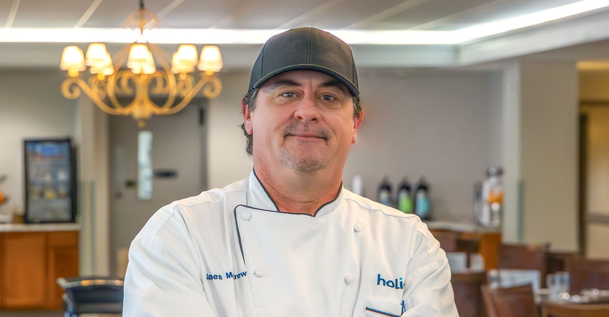 Chef wearing a white jacket and black cap stands confidently in a dining room with a chandelier and chairs in the background.