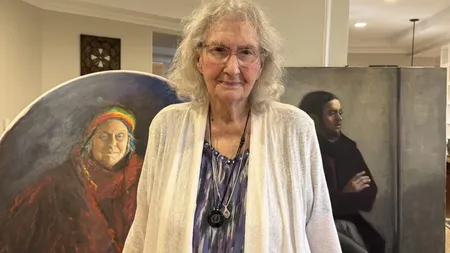 Older woman in glasses and white cardigan stands in front of two painted portraits, smiling indoors.