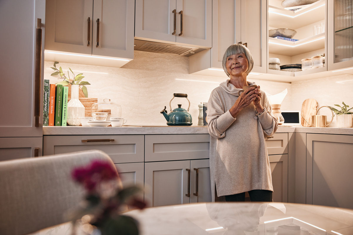 Senior woman in a cozy kitchen holding a mug, surrounded by warm lighting, plants, and kitchenware on the counter.