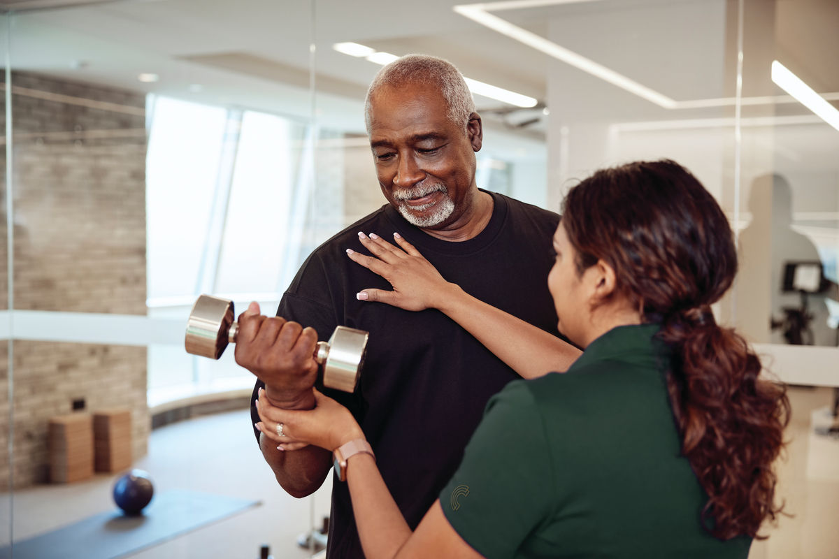 Man lifting a dumbbell with assistance from a trainer in a gym, both smiling. Bright, modern interior with exercise equipment visible.