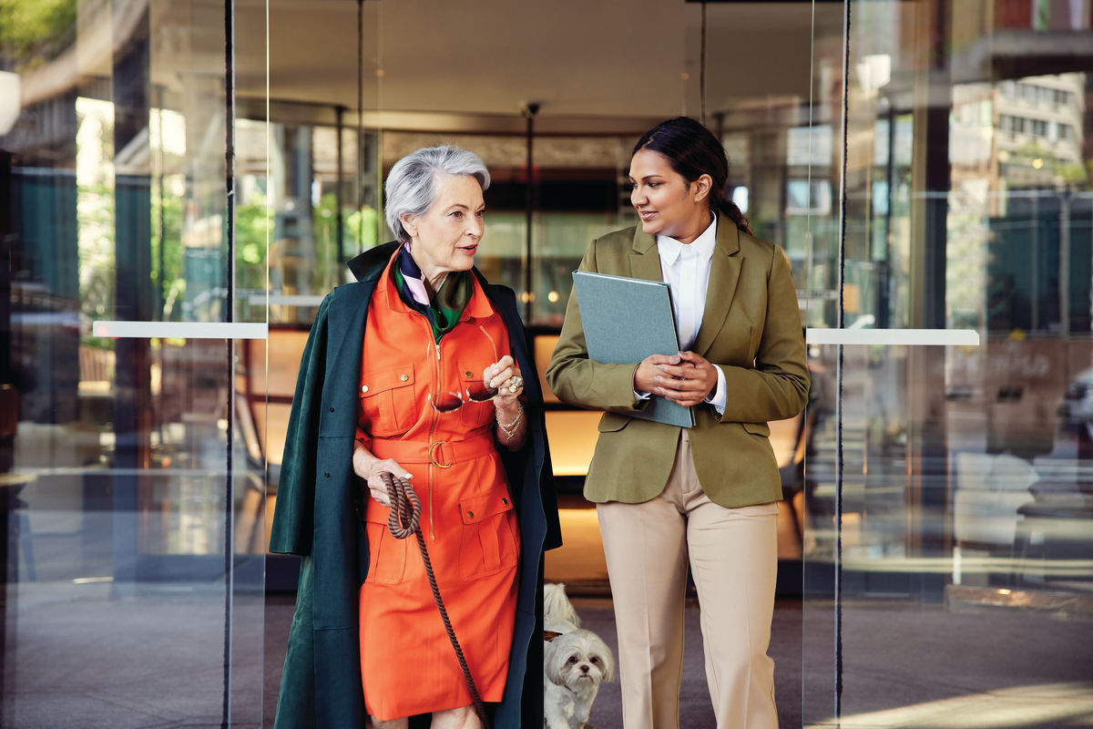 Two women walking and conversing outside a building; one in an orange coat, the other in a green suit holding a folder, with a small dog nearby.