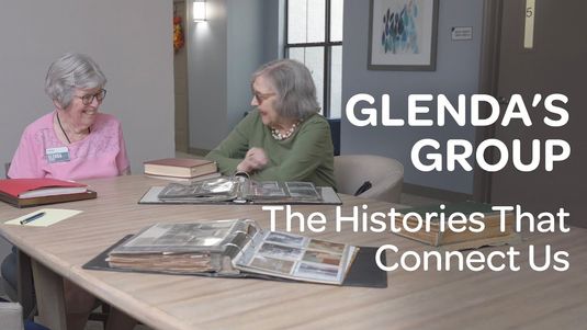 Two senior women sit at a table with photo albums, smiling and talking. Text reads "GLENDA'S GROUP: The Histories That Connect Us."