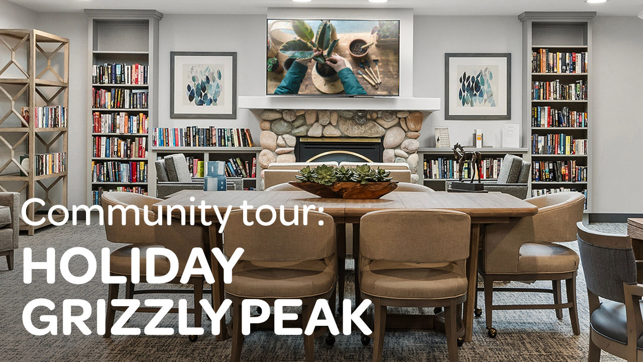Cozy library with a stone fireplace, bookshelves, and a large wooden table surrounded by beige chairs. Text reads "Community tour: Holiday Grizzly Peak."