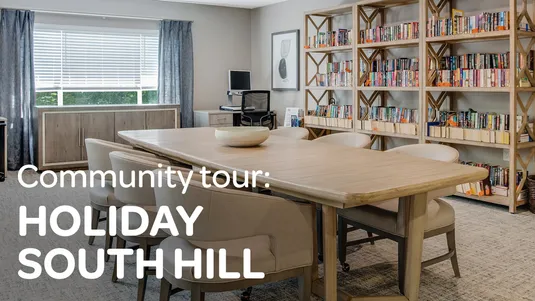 A community room with a table, chairs, bookshelves, a computer desk, and framed art. Text overlay: "Community tour: Holiday South Hill."