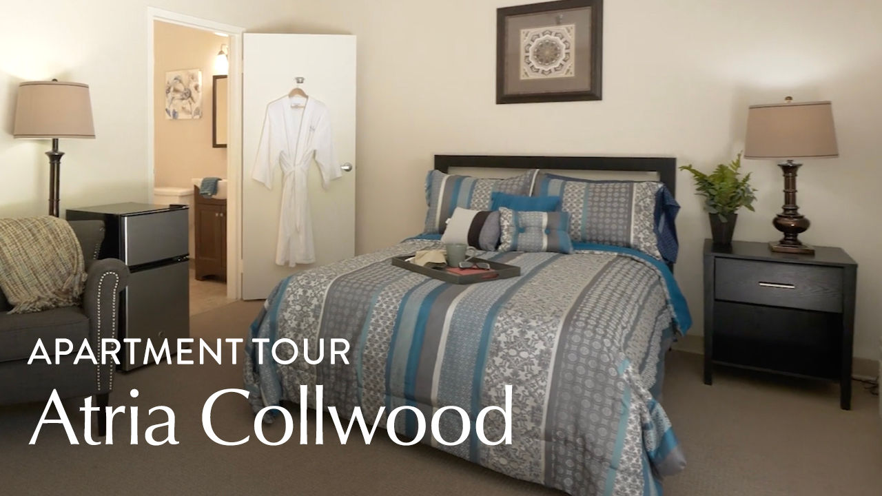 A cozy bedroom with a neatly made bed, decorative pillows, a robe on the door, and a side table with a lamp. Text reads "Apartment Tour Atria Collwood."