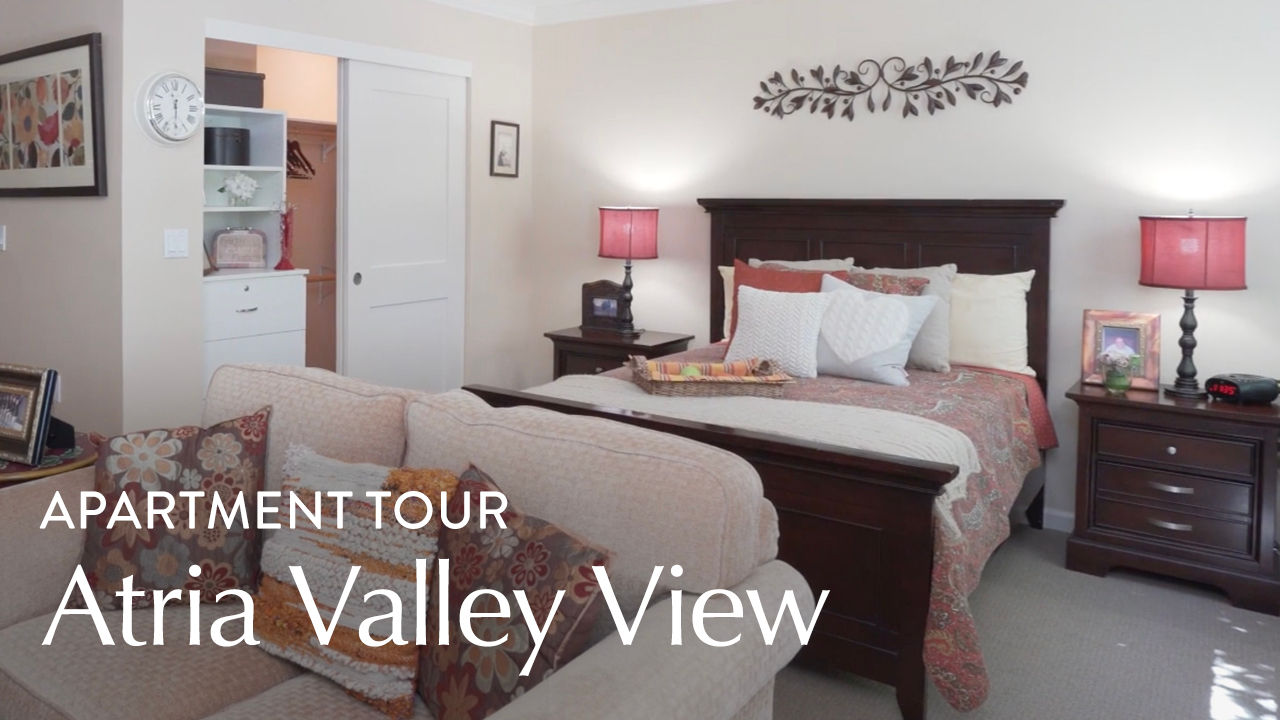 Cozy bedroom with a wooden bed, patterned bedding, red lamps, and a small living area. Text reads "Apartment Tour Atria Valley View."