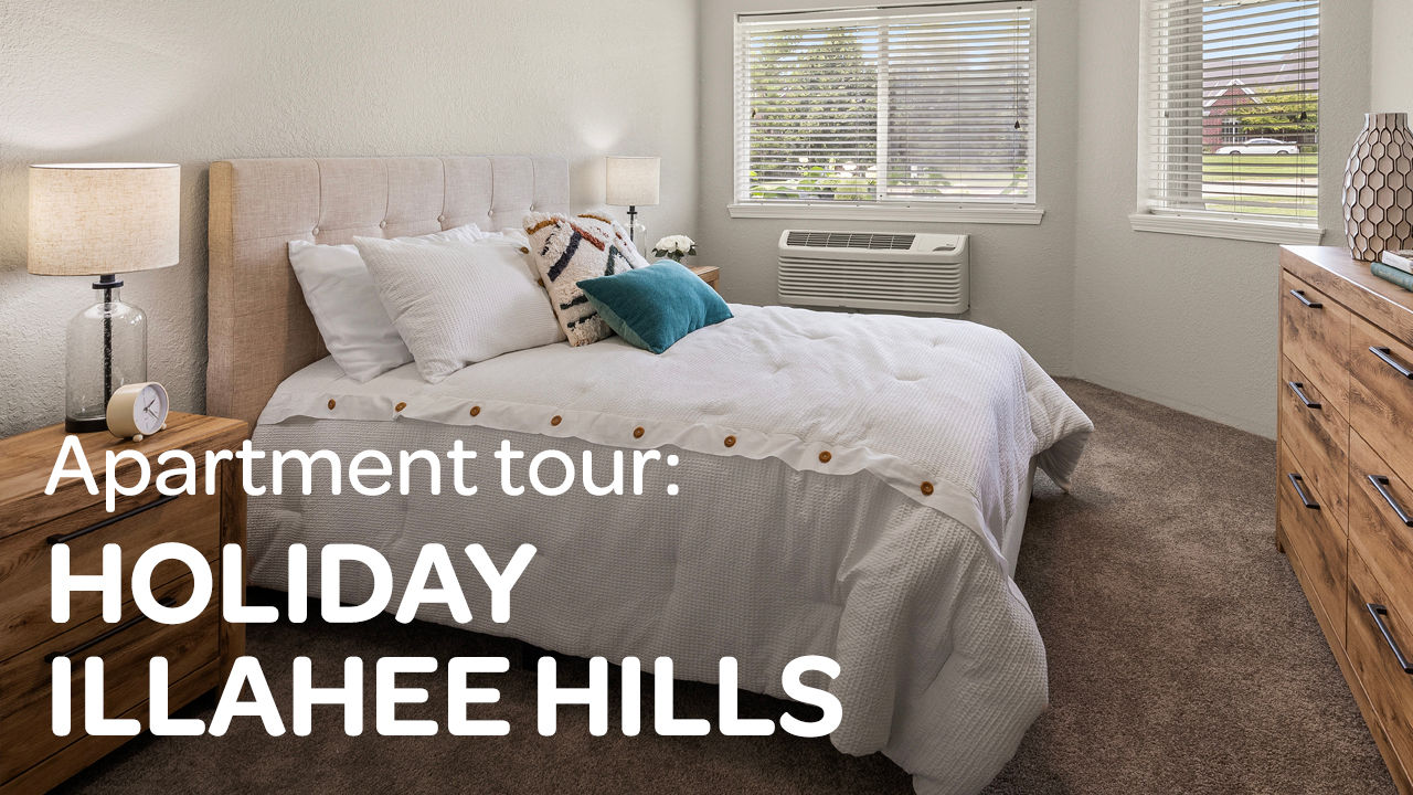 Cozy bedroom with a neatly made bed, bedside lamps, and a window with blinds. Text overlay: "Apartment tour: Holiday Illahee Hills."