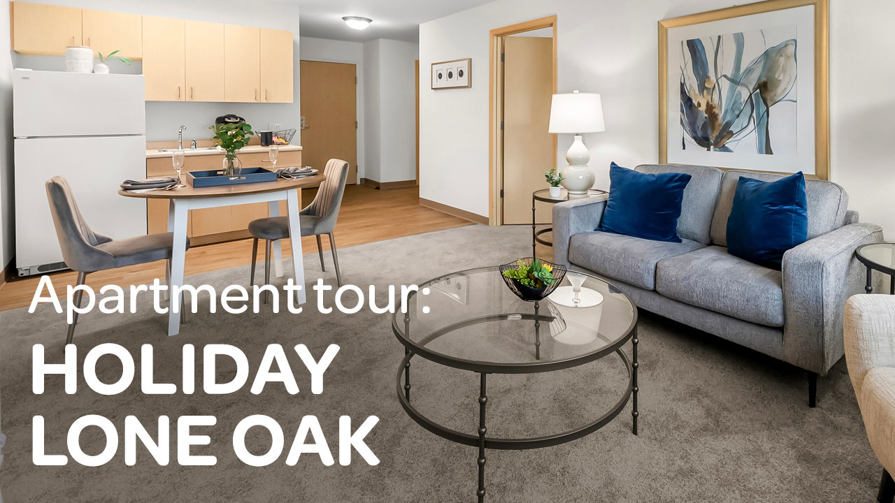 Modern apartment living room with gray sofa, glass coffee table, dining area, and kitchen. Text reads "Apartment tour: Holiday Lone Oak."