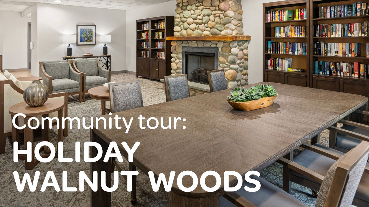 Cozy room with a large wooden table, chairs, stone fireplace, bookshelves, and seating area. Text: "Community tour: HOLIDAY WALNUT WOODS."
