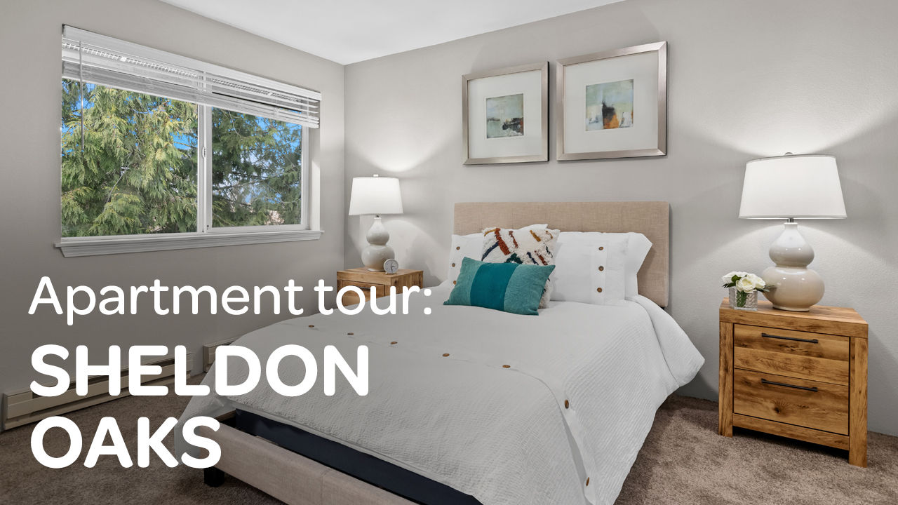 Bedroom with a large bed, white bedding, two wooden nightstands, lamps, and wall art. Window shows green trees. Text reads "Apartment tour: SHELDON OAKS."