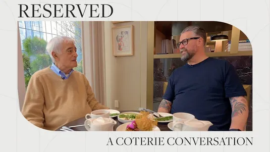Two men seated at a table engaged in conversation, with food and drinks in front of them, set in a cozy room.