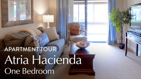 Cozy living room with a sofa, armchair, coffee table, and lamp. Large window with curtains. Text: "Apartment Tour Atria Hacienda One Bedroom."