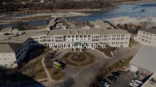Aerial view of a large building labeled "Atria on Roslyn Harbor," surrounded by parking lots, a roundabout, and a river in the background.
