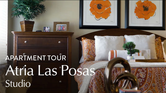Cozy studio bedroom with a wooden dresser, floral bedding, and framed flower art. Text reads "Apartment Tour Atria Las Posas Studio."