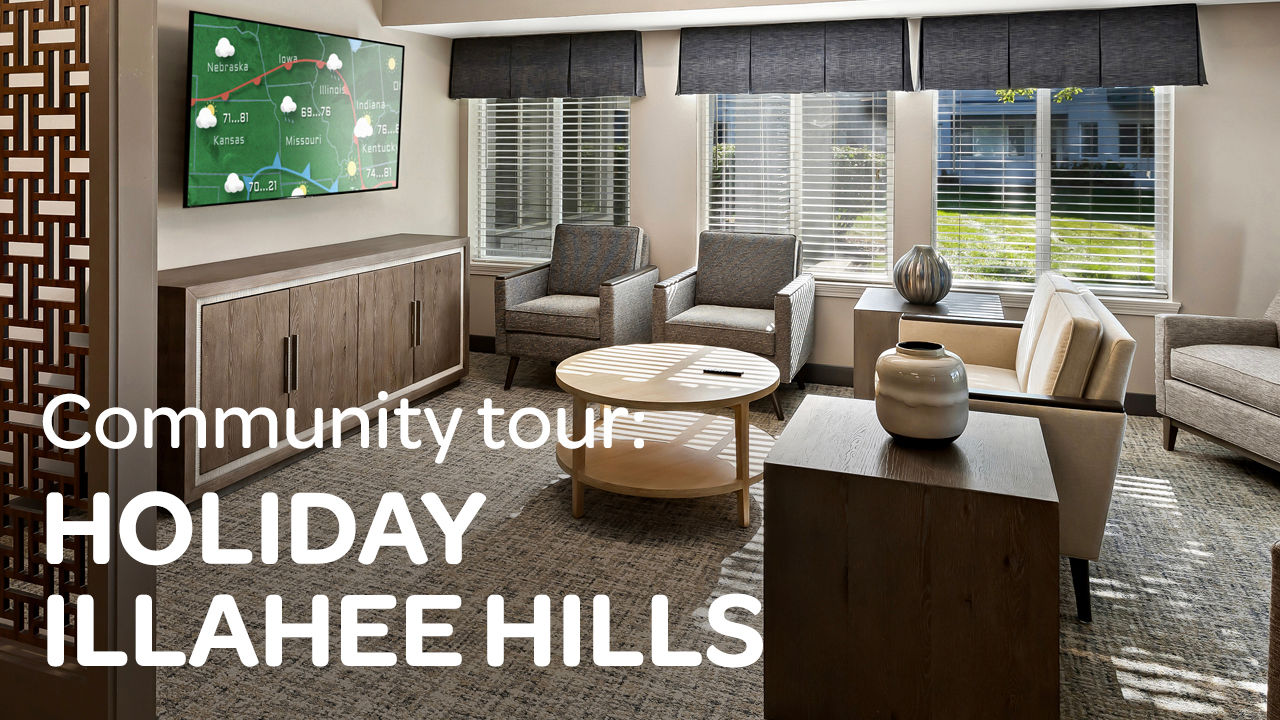 Modern lounge with chairs, round table, decorative vase, and wall-mounted TV displaying a community tour at Holiday Illahee Hills.