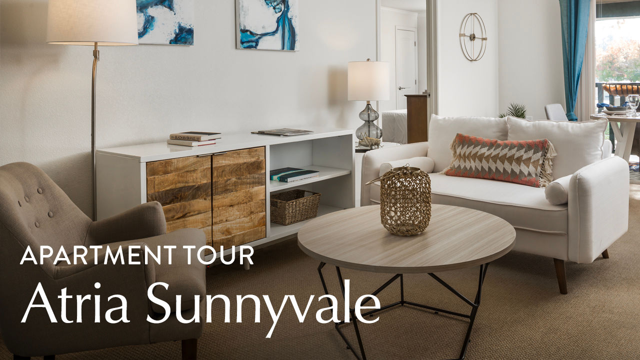 Modern living room with a white sofa, gray armchair, wooden coffee table, and a sideboard. Text reads "Apartment Tour Atria Sunnyvale."