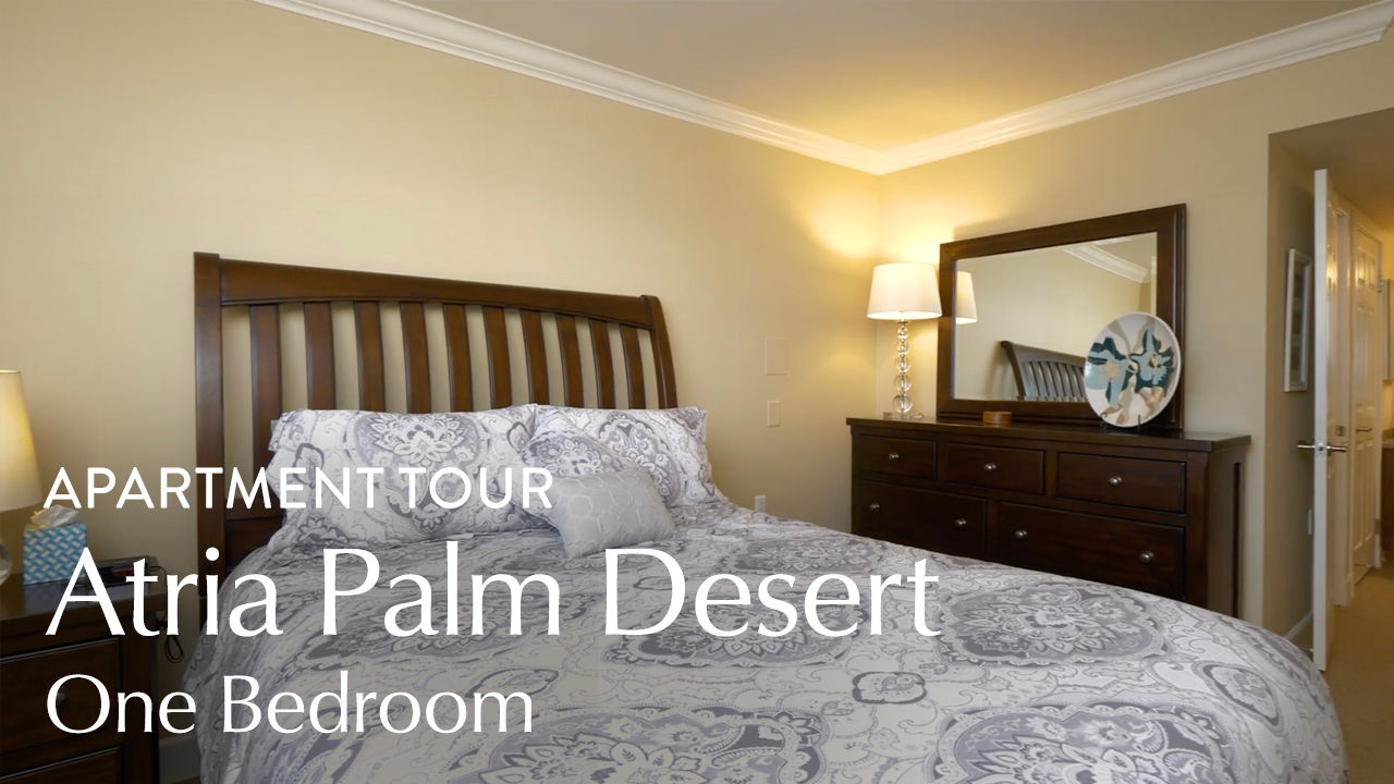 Cozy bedroom with a wooden bed, patterned bedding, dresser, mirror, and a lamp, labeled "Apartment Tour: Atria Palm Desert One Bedroom."