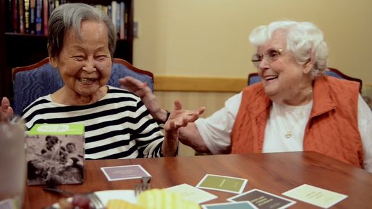 Two older adult ladies laugh together while seated over a game of storytelling at Atria.