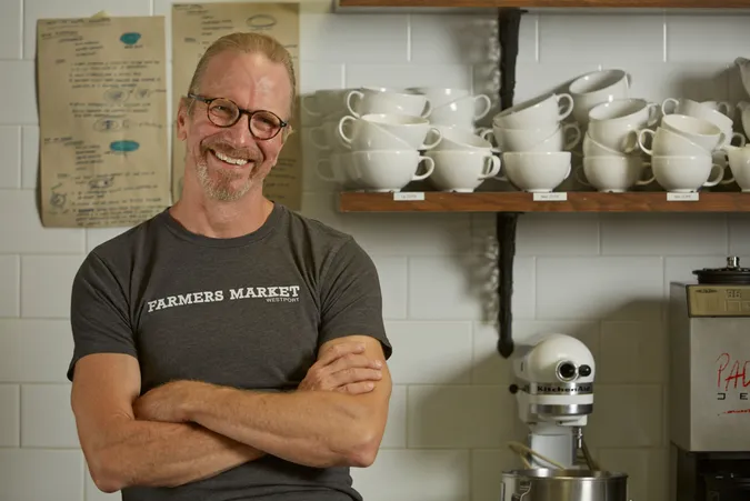 Smiling man with glasses and a "Farmers Market" shirt stands in a kitchen with stacked cups and a mixer on shelves behind him.