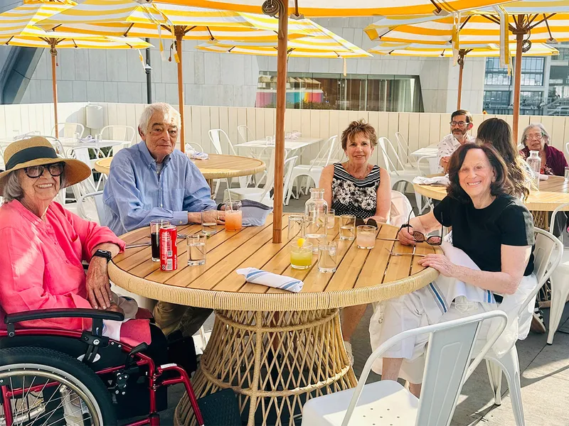 A group of senior living residents sitting at a round outdoor table under striped umbrellas, enjoying drinks and smiling.