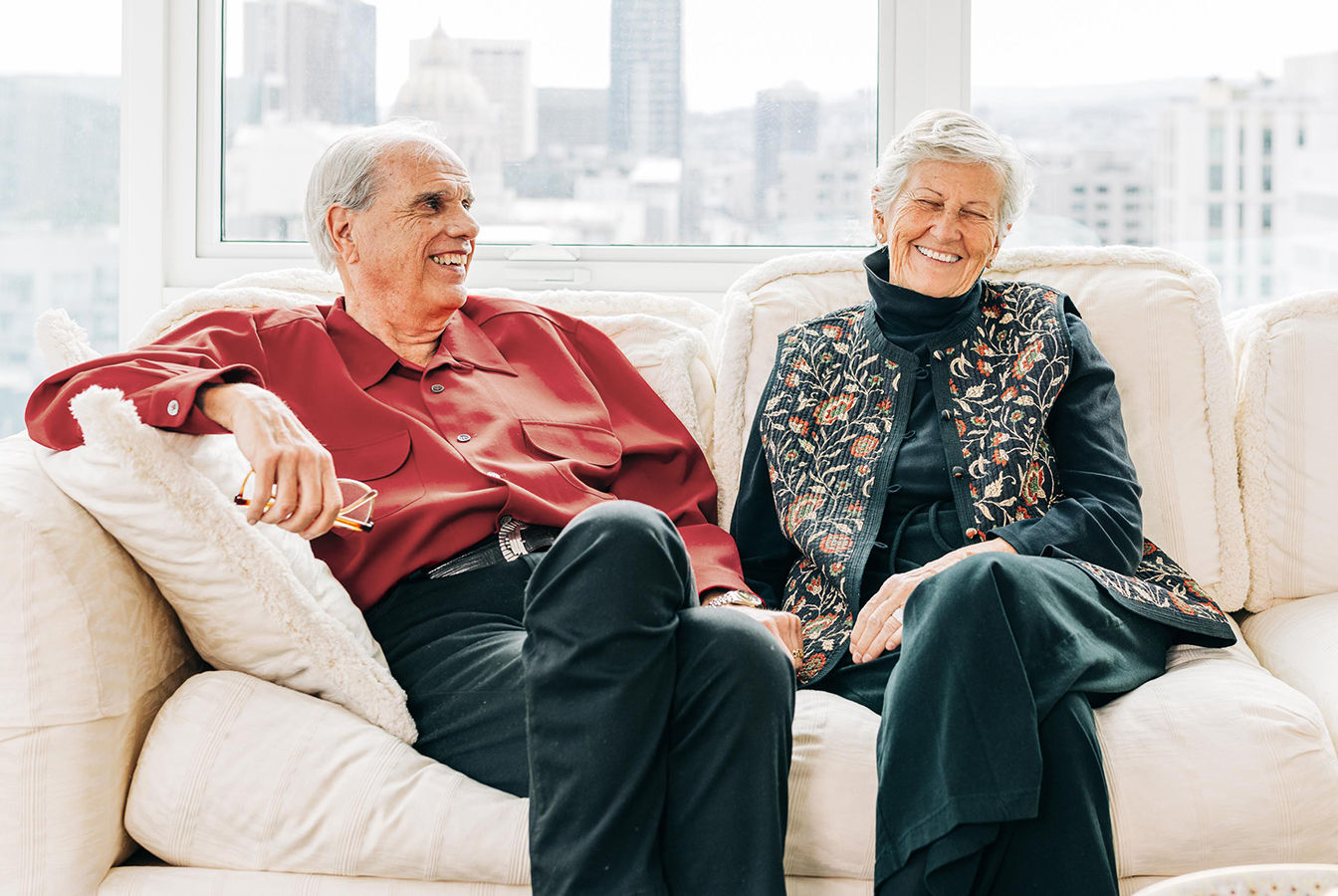 Older couple smiling and sitting on a sofa in a bright living room with cityscape background.
