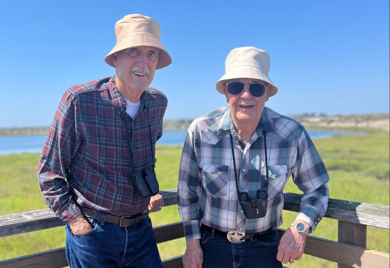 Two senior men in plaid shirts and bucket hats stand on a wooden deck by a grassy landscape, each with binoculars around their necks.