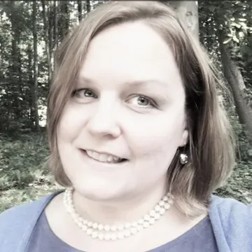 Smiling woman with short hair and pearl necklace, standing in a forested area.
