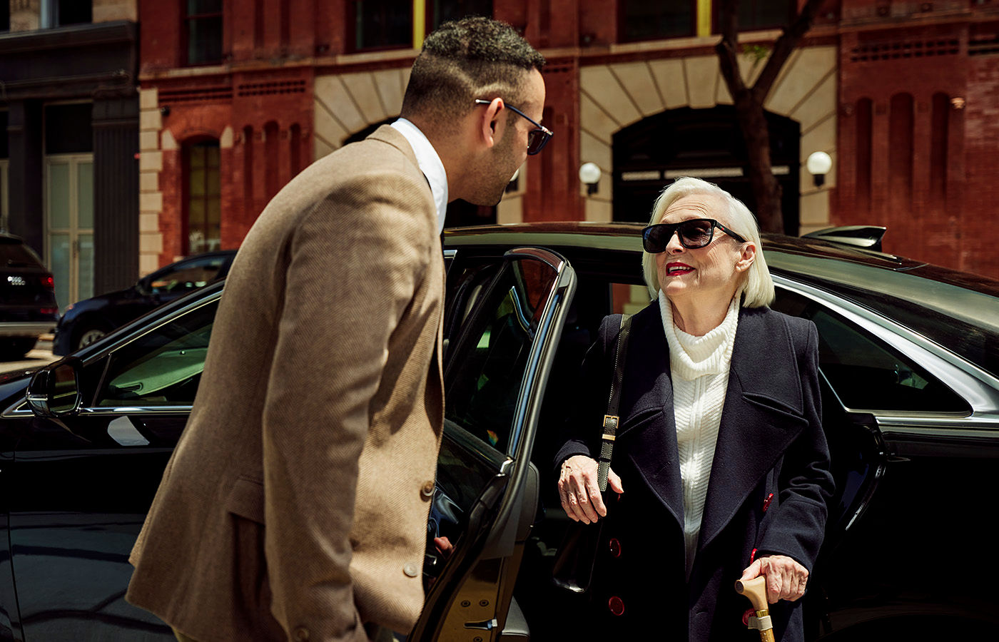 Senior woman exiting a car downtown wearing sunglasses and a peacoat. Woman is being helped by gentleman in suit and sunglasses.