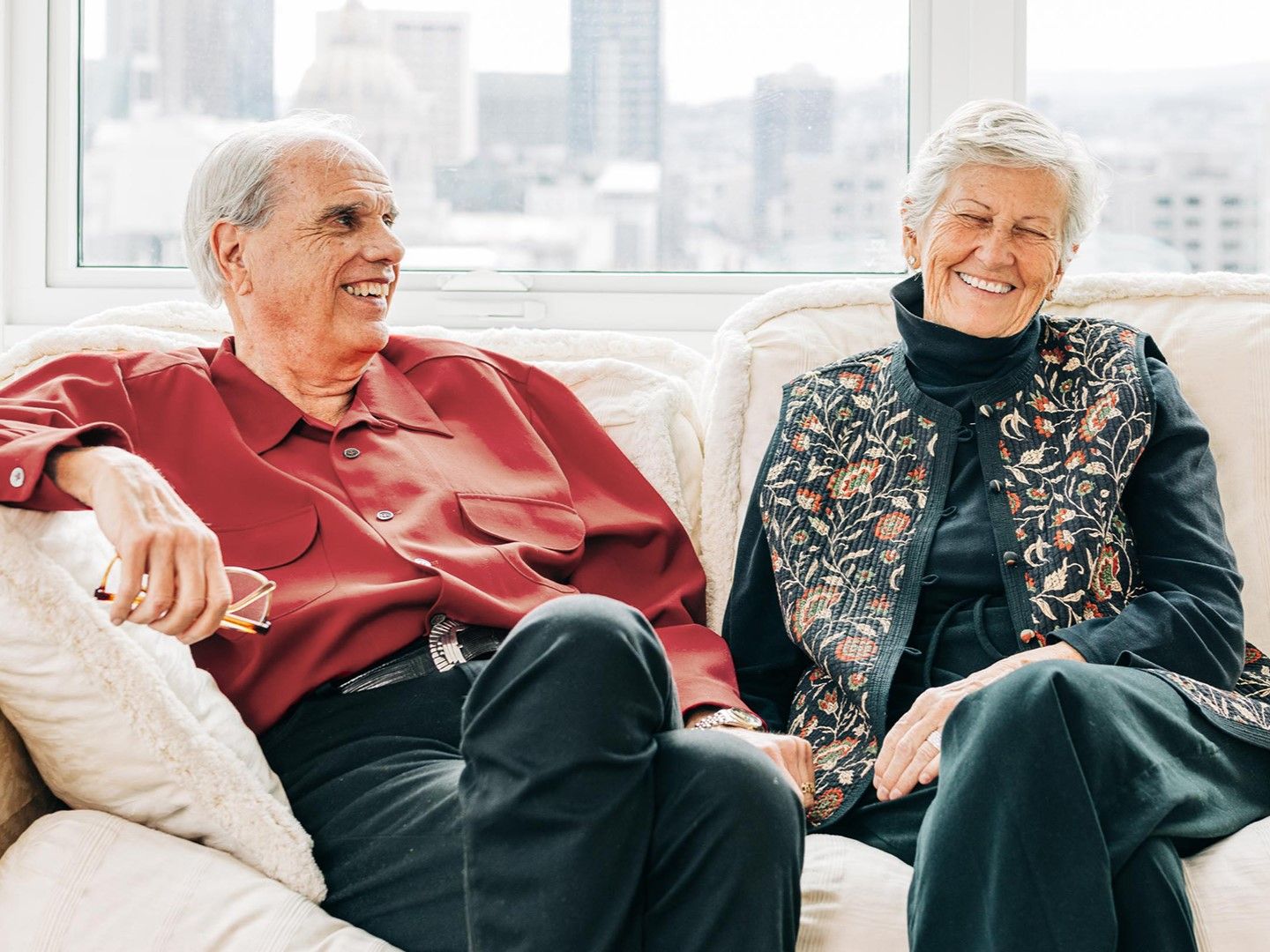 Older couple smiling while sitting on a cozy white sofa in a bright room with city views.