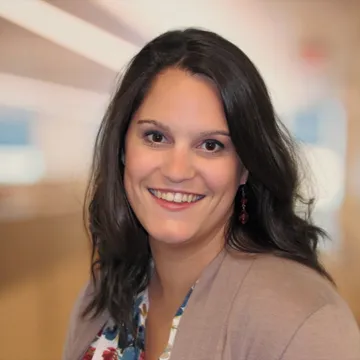 Smiling woman with long dark hair wearing a floral shirt and beige cardigan, in a softly blurred interior setting.