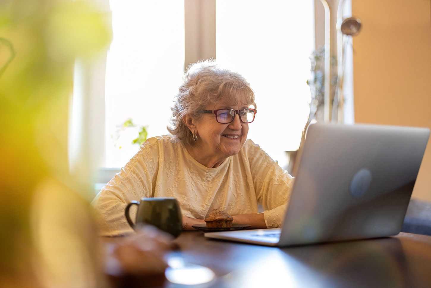 Senior woman sitting at kitchen table with a cup of coffee smiling at a laptop.