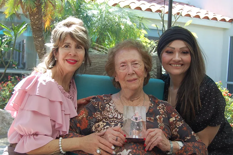 Three women smiling together outdoors; the older woman holds a photograph.