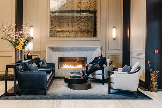 Older person sitting in a modern living room with a lit fireplace, surrounded by elegant furniture and floral arrangements.
