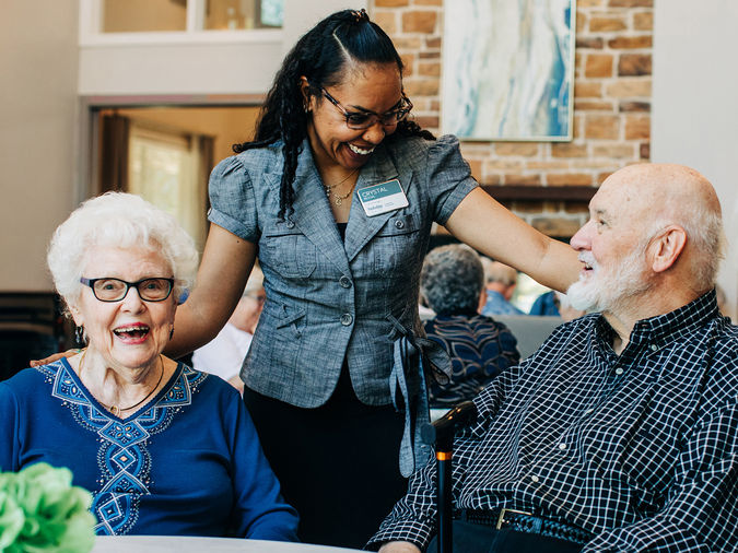 A smiling woman stands between two seniors seated at a table, engaging in conversation in a bright room.