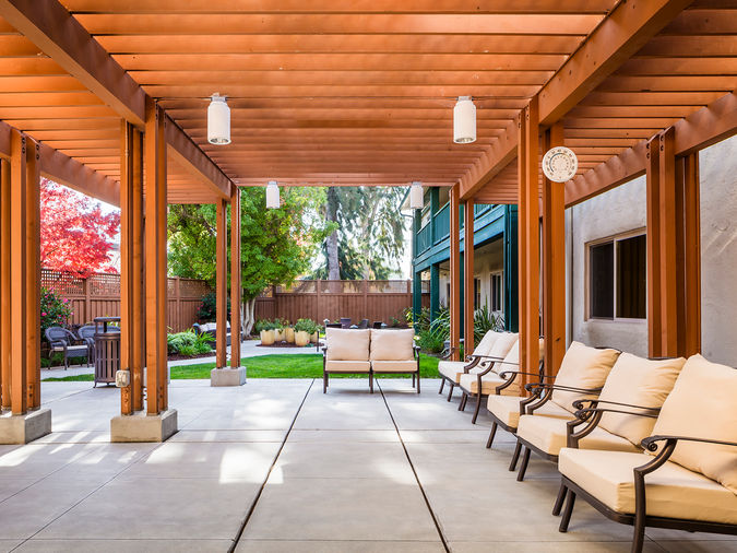 Outdoor patio with wooden pergola, cushioned seating, and a view of a lush garden with trees and potted plants.