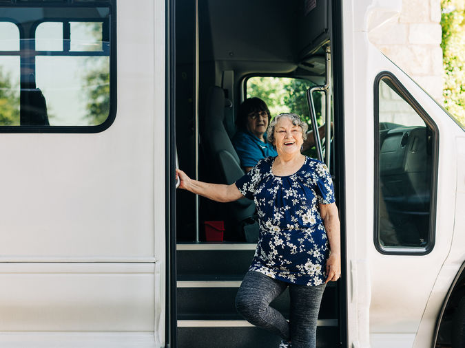 Senior woman smiling at the door of a bus, with another person seated inside. The bus is parked, and greenery is visible in the background.