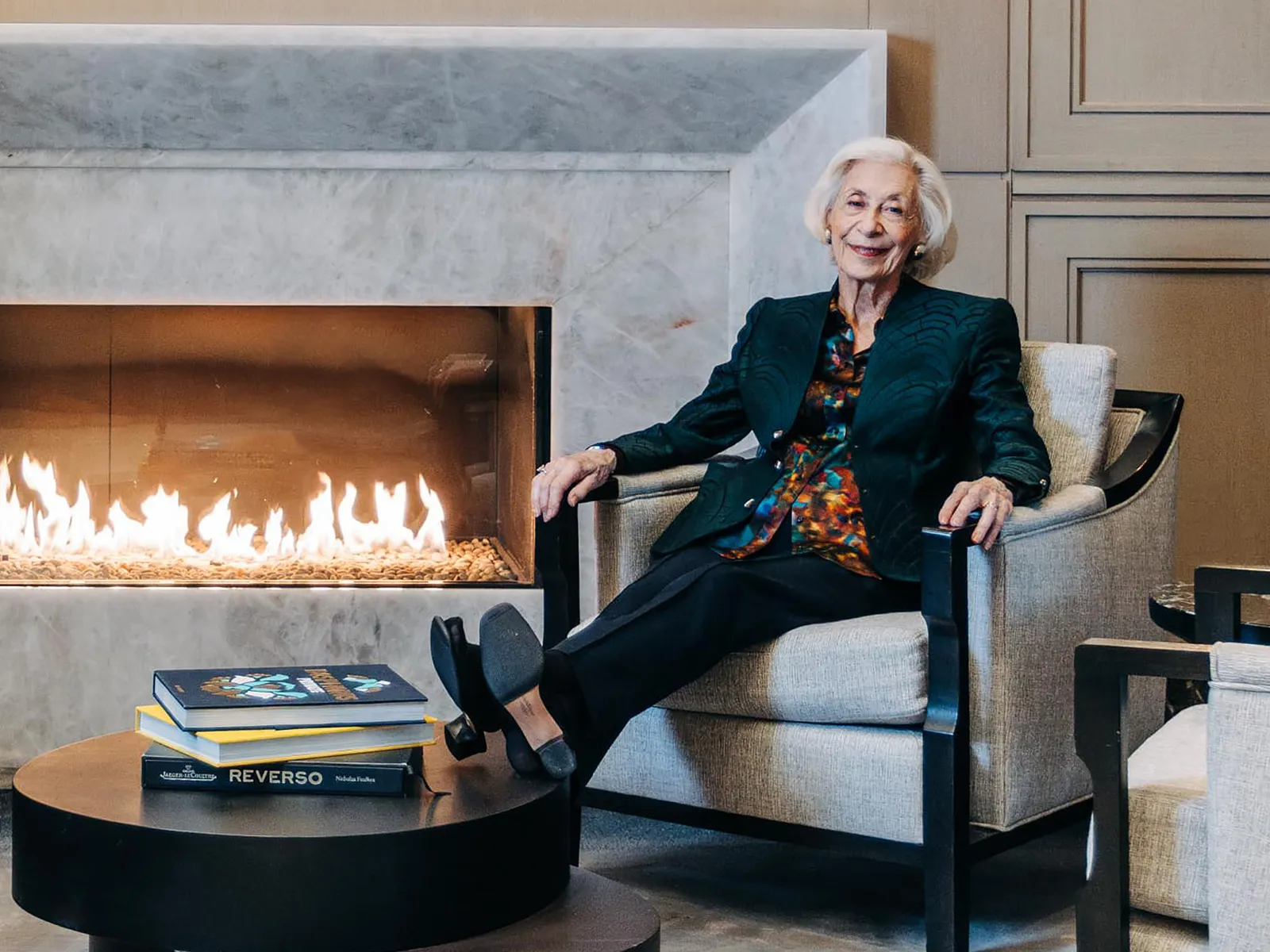 Older woman sitting comfortably by a modern fireplace, smiling, with books on a round table in a cozy, elegant room.