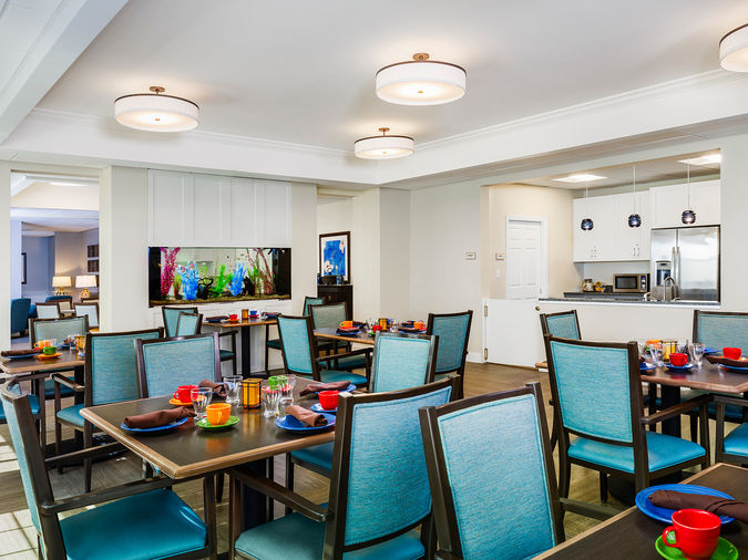 A modern dining room with multiple tables set with colorful plates and cups, teal chairs, and bright lighting. A kitchen is visible in the background.
