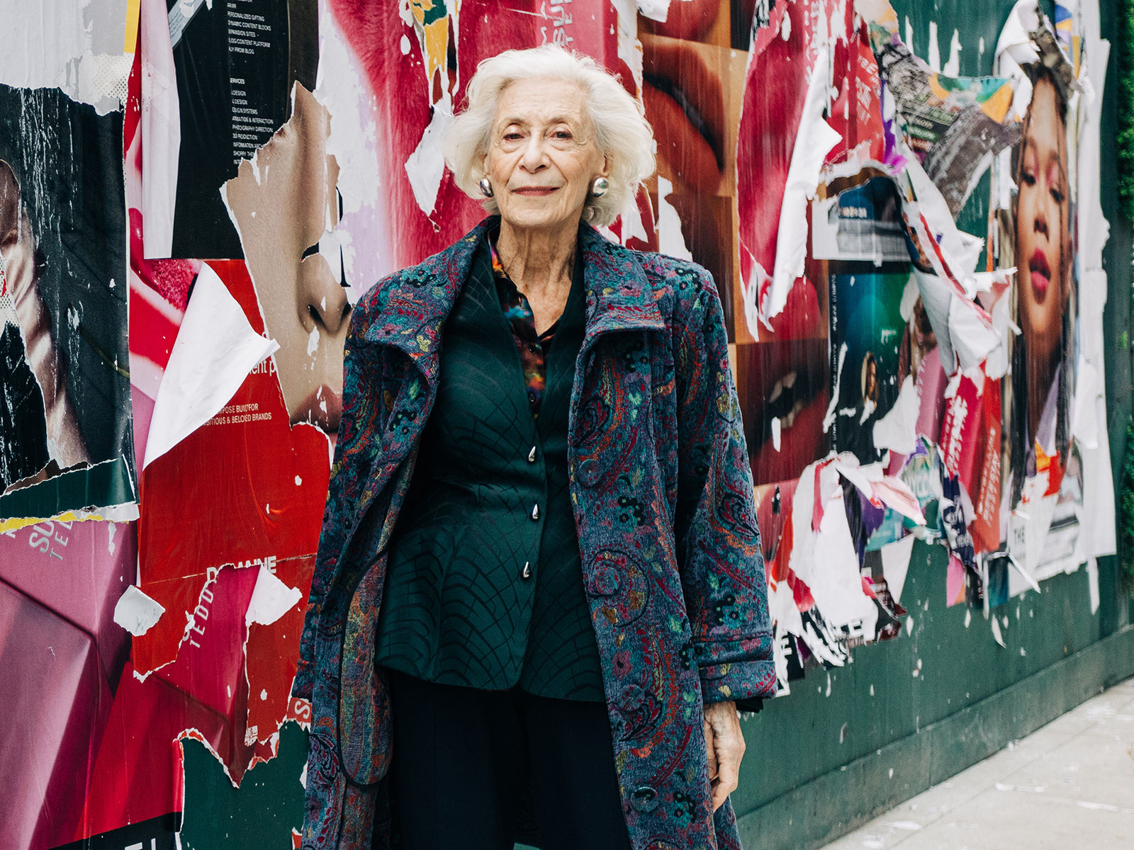 Older woman with white hair in a patterned coat stands in front of a wall covered with torn, colorful posters.