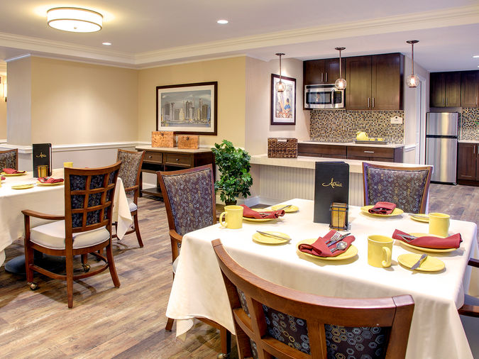 A cozy dining area with elegantly set tables, wooden chairs, warm lighting, and a modern kitchen in the background.