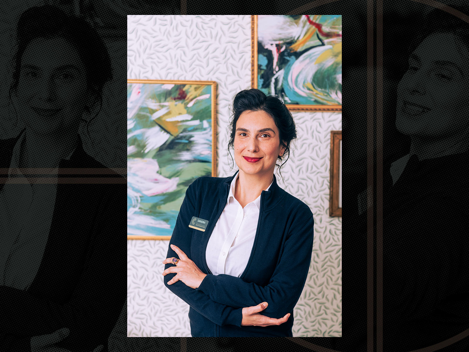 Woman in a navy blazer stands smiling with arms crossed. Abstract paintings hang on a patterned wall in the background.