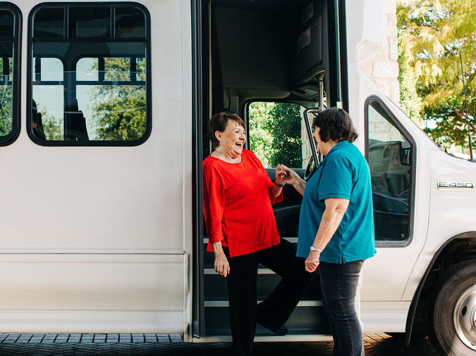 Two women smiling and talking at the entrance of a white shuttle bus, with one stepping onto it and the other standing outside.