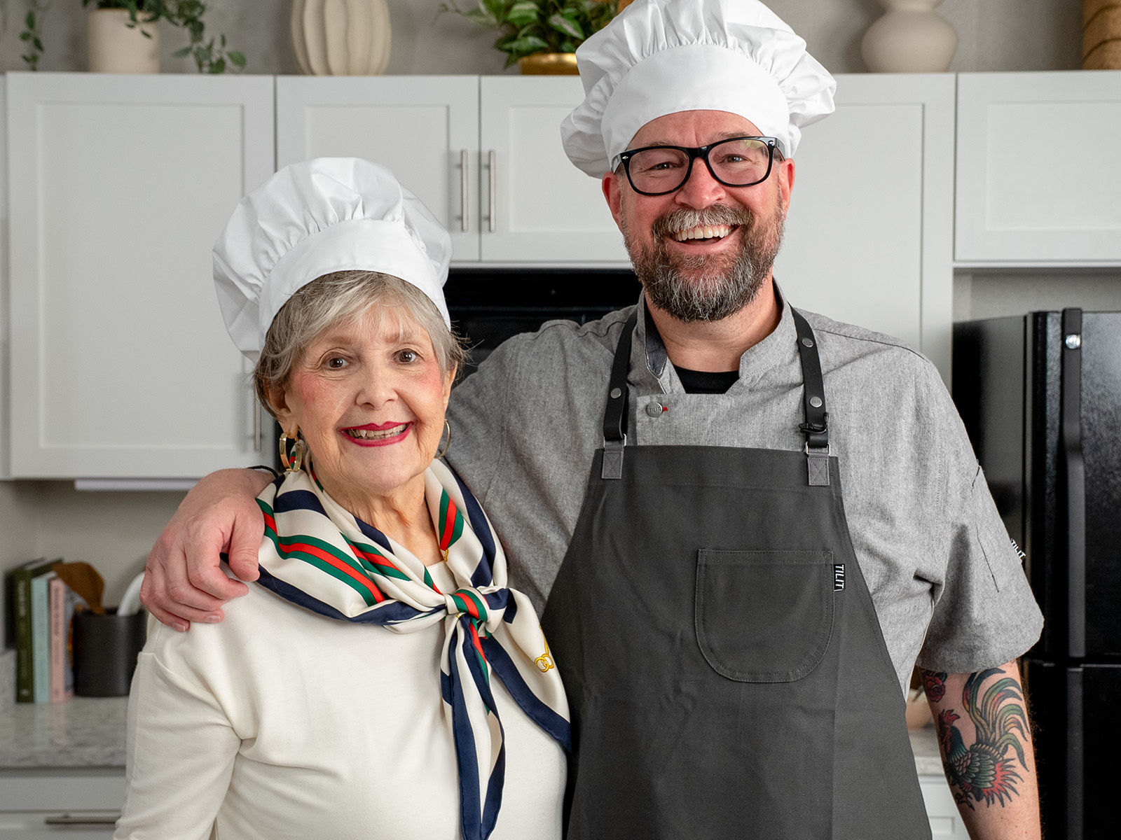 Two smiling chefs in a kitchen, one older woman wearing a scarf and chef hat, and a bearded man in an apron and chef hat.