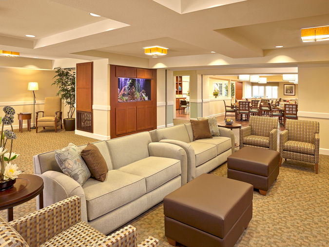 Spacious lounge with beige sofas, patterned chairs, and a fish tank. Warm lighting and a dining area in the background create a cozy atmosphere.