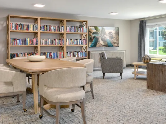 A cozy library room with a wooden table surrounded by beige chairs, bookshelves filled with books, a wall-mounted TV, and a large window with curtains.