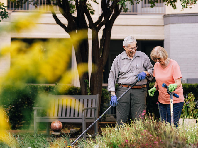 Senior couple gardening together, wearing gloves, surrounded by greenery and flowers, with a bench and tree in the background.